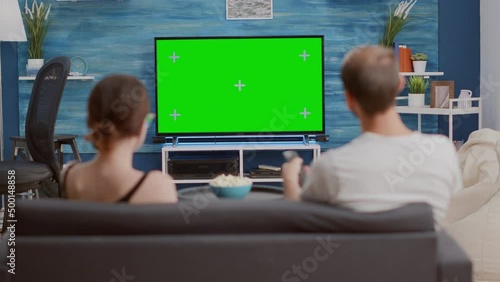 Couple sitting on couch watching television on green screen flat tv talking about favourite show and zapping in modern living room. Man and woman having popcorn enjoying movie on chroma key display.