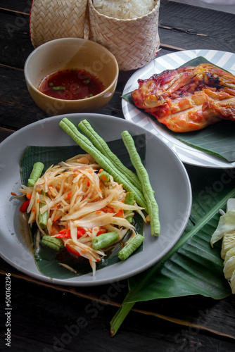 Thai food dish both in Thailand and Asia, "Papaya Salad" or as we call it "Somtum" is complemented with grilled chicken and sticky rice with fresh stir-fries. Served on the black wooden table.