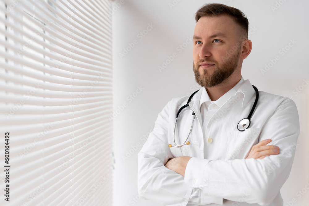 Portrait of a confident male doctor with crossed arms standing in a clinic near a window with jalousie. Portrait of a confident male doctor with crossed arms standing in a clinic near a window with jalousie.