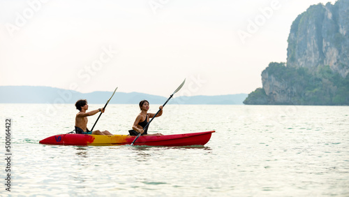 Young Asian man and woman kayaking together in the sea at tropical lagoon island at summer sunset. Male and female friends enjoy outdoor activity lifestyle and water sports on beach holiday vacation