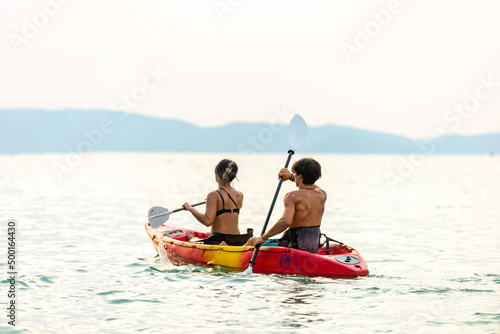 Young Asian man and woman kayaking together in the sea at tropical lagoon island at summer sunset. Male and female friends enjoy outdoor activity lifestyle and water sports on beach holiday vacation