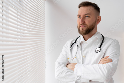 Portrait of a confident male doctor with crossed arms standing in a clinic near a window with jalousie.