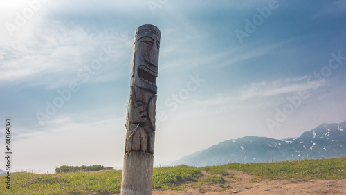 There is an old weathered wooden idol in the valley. The totem in the form of a pillar is decorated with carvings. Mountains against a background of blue sky and clouds. Kamchatka. Vilyuchinsky pass. photo
