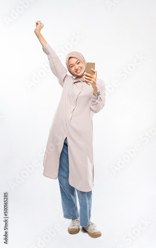 Young asian woman making a call using a cell phone and showing excited gesture with arm raised on isolated background