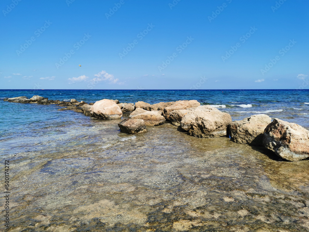 The coast of the Mediterranean Sea, waves, clear water, a stone ridge against a blue sky with clouds.