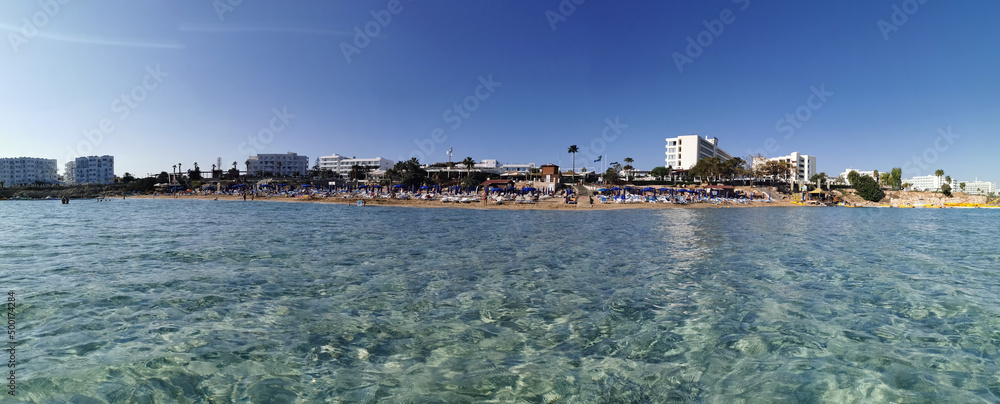 Protaras. Famagusta area. Cyprus. Panorama of Fig Tree Bay beach, people sunbathing and swimming, hotel buildings behind the beach against the blue sky. View from the sea.