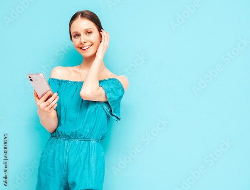 Young beautiful smiling female in trendy summer blue overalls. Sexy carefree woman posing near wall in studio. Positive brunette model looking at cellphone screen. Holding smartphone and using apps