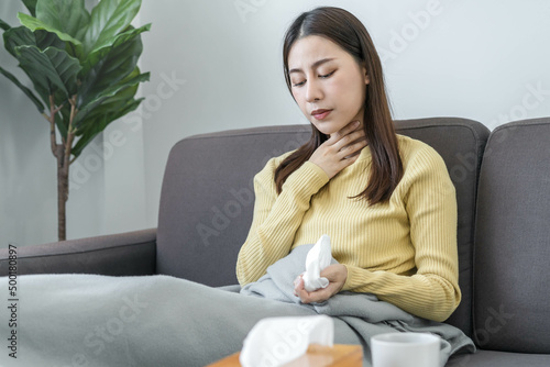 Sick, hurt or pain asian young woman, girl with sore throat, headache have a fever, flu and use tissues paper sneezing nose, runny sitting on sofa bed at home. Health care person on virus seasonal. © KMPZZZ