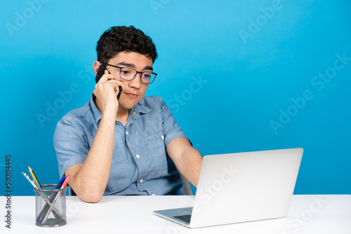 Hispanic young man talking on phone in front of laptop isolated on blue background.