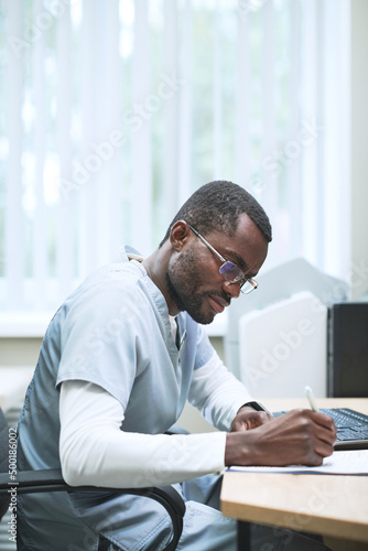 Concentrated African-American male doctor in eyeglasses sitting at desk and filling medical card in office