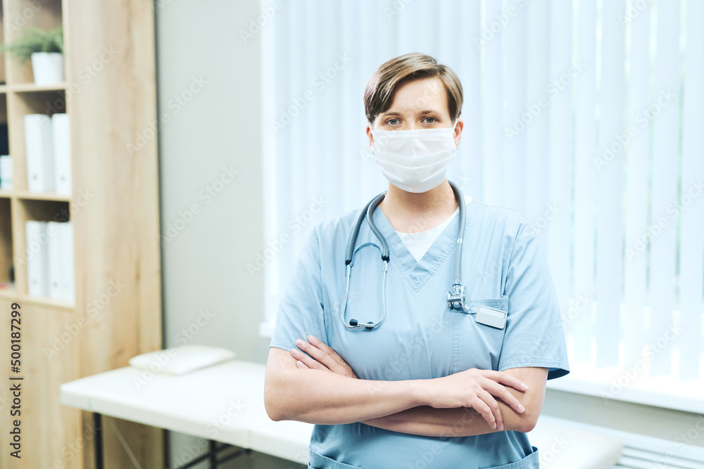 Portrait of fenale medical intern in facial mask standing with crossed arms against examination couch in hospital Portrait of fenale medical intern in facial mask standing with crossed arms against examination couch in hospital
