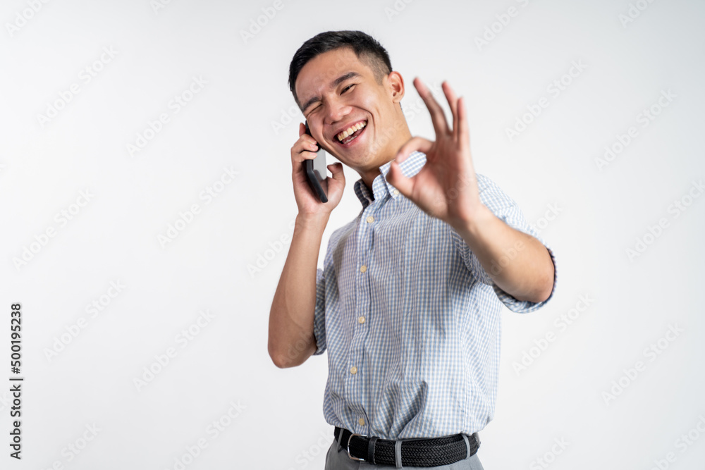 Happy asian young man showing okay gesture while making phone call on isolated background