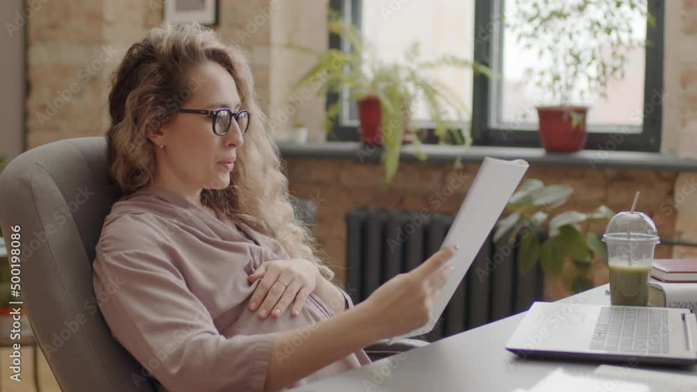 Medium side-view of long-haired curly expecting woman wearing eyeglasses, sitting at desk in modern office at daytime, reading financial report Medium side-view of long-haired curly expecting woman wearing eyeglasses, sitting at desk in modern office at daytime, reading financial report