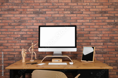 Modern computer and tablet with blank screens on desk near brick wall, space for design. Comfortable workplace