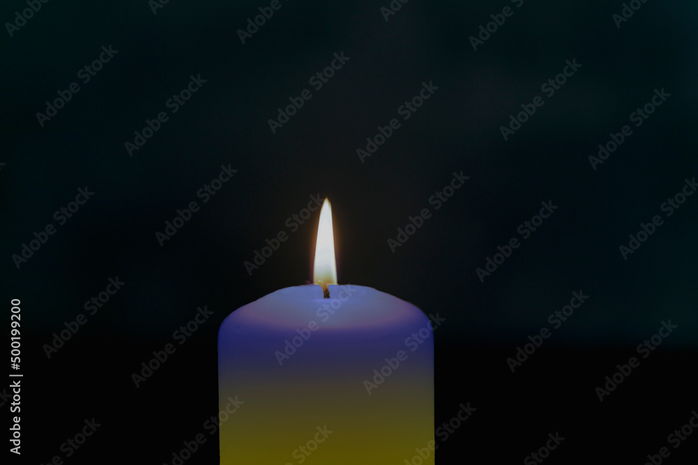Płomień świecy w ciemnościach. Świeca ma zabarwienie niebiesko żółte, symbolizuje solidarność z Ukrainą. - obrazy, fototapety, plakaty 