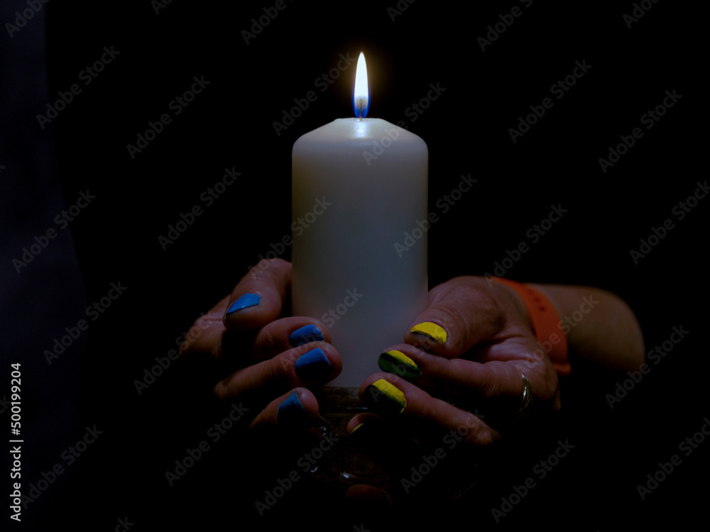 Płomień białej świecy w ciemności trzymanej przez kobietą. Paznokcie kobiety pomalowane są na niebieski i żółty kolor. - obrazy, fototapety, plakaty 