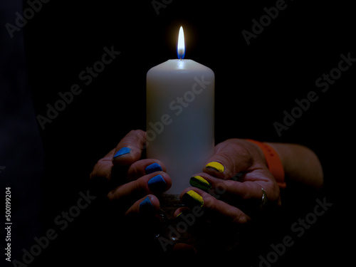 Płomień białej świecy w ciemności trzymanej przez kobietą. Paznokcie kobiety pomalowane są na niebieski i żółty kolor.