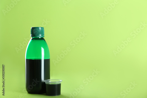 Bottle of cough syrup and measuring cup on light green background. Space for text