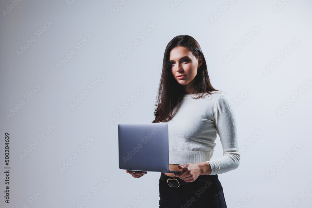 Young woman manager in a white shirt holds a portable laptop in her hands while standing on a gray background Young woman manager in a white shirt holds a portable laptop in her hands while standing on a gray background