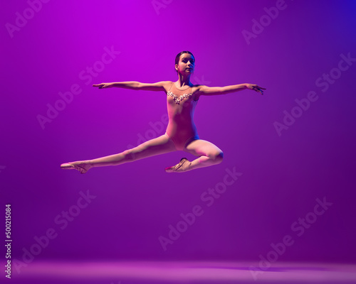 Portrait of young little ballet dancer  teen jumping isolated on purple background in neon light. Art  grace  beauty  ballet school concept