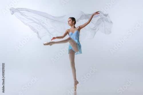 Beautiful young girl, aspiring ballerina in blue dress and pointes dancing with cloth isolated on gray background. Art, grace, beauty, ballet school concept