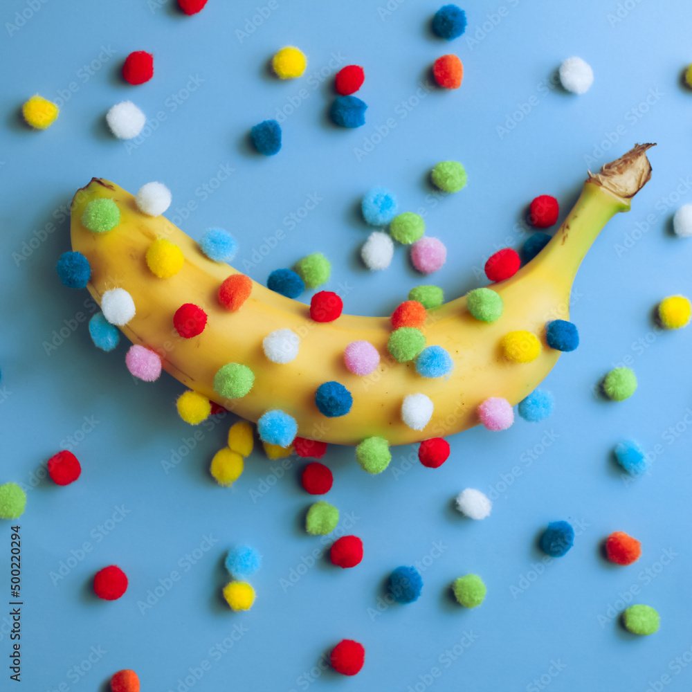 Funny banana with pom-poms, a banana covered with round colored pom-poms on a blue background and surrounded by scattered pom-poms, strange fruit concept