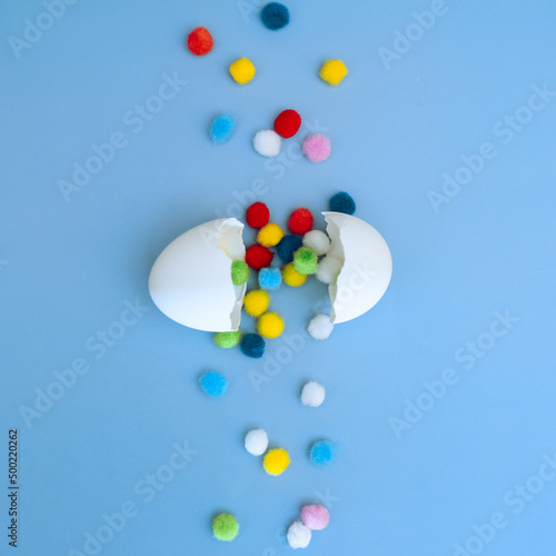 Cracked white egg with multi-colored round pom-poms falling out of it on a blue background  top view