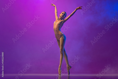 Beautiful flexible young female ballet dancer, teen in stage outfit and pointes dancing isolated on purple background in neon light with smoke. Art, grace, beauty