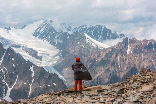 Hiker at very high altitude with view to long glacier tongue and high snow mountains under cloudy sky. Man on windy stone hill against huge mountains with low clouds. Man and majestic mountains nature