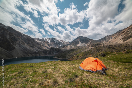 Vivid orange tent on grassy hill with view to mountain lake of phantom blue color among high mountains in changeable weather. Tent near deep blue lake among sunlit large mountains under cloudy sky.