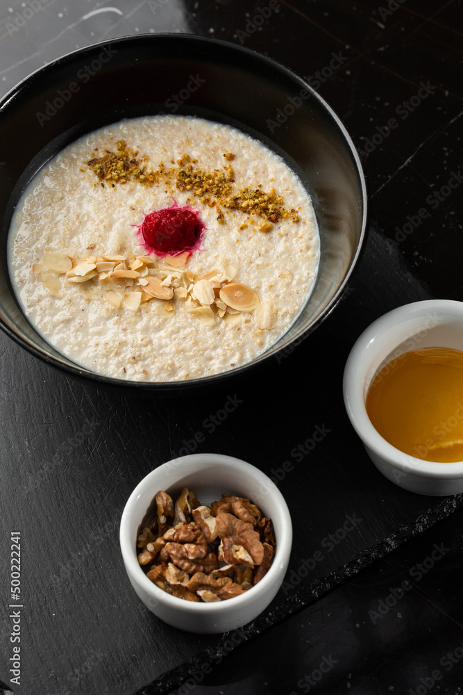 Semolina porridge for breakfast oat with nuts in black bowl served with honey isolated on black stone background. Homemade food. Tasty breakfast. Semolina porridge for breakfast oat with nuts in black bowl served with honey isolated on black stone background. Homemade food. Tasty breakfast.
