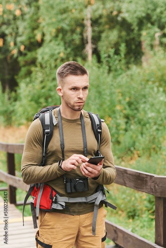 Pensive young hiker with stubble using smartphone while standing on bridge outdoors