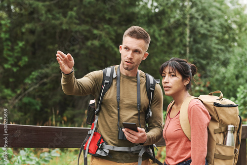 Young Caucasian hiker with camera hanging on neck gesturing hand and using phone while explaining route to woman © Mediaphotos