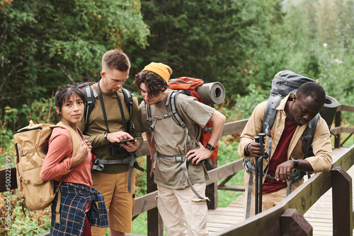 Group of young multi-ethnic hikers with backpacks standing on bridge in forest and analyzing route on phone