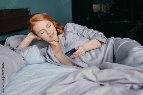 young beautiful red-haired woman dressed in blue pajamas lies on the bed and uses a smartphone