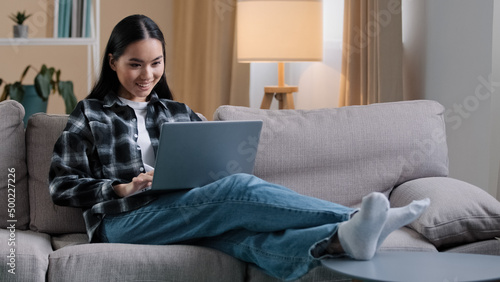 Carefree Asian girl enters living room sit down on comfortable sofa takes laptop with smile and watches video online woman uses computer sitting on couch at home casual browsing net resting typing