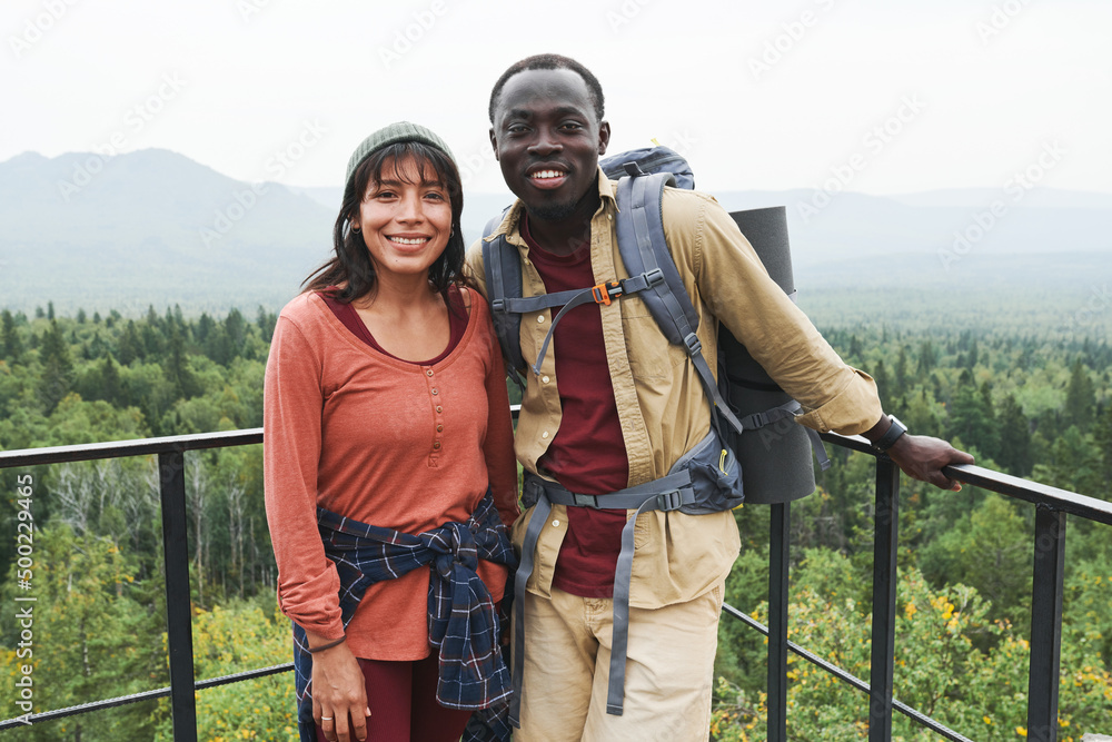 Fototapeta premium Happy multi-ethnic couple standing at railings against forest landscape and looking at camera at hike