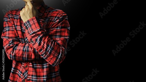 An entrepreneur in a red and black plaid shirt puts his hand on his chin making contemplative thoughtful and planning gestures on a black background.