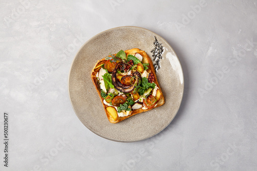 Bruschetta with octopus, vegetables and sauce on gray ceramic plate. Toast with octopus in contemporary style. Chef concept bruschetta in modern ceramic dishware. Handmade dinner plate.