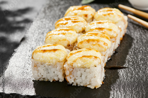 Baked Maki sushi on dark slate. Hot cheese maki with unagi sauce. Sushi roll with baked cheese and unagi sauce topped. Style concept japanese menu with black background, leaves and hard shadow.