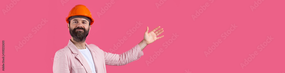 Portrait of young construction engineer wear orange hard hat, in a pink jacket standing on red studio background. The man points with his hand. Portrait of young construction engineer wear orange hard hat, in a pink jacket standing on red studio background. The man points with his hand.