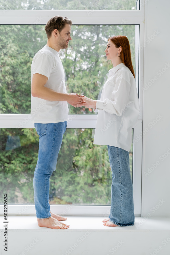 Side view of positive barefoot couple holding hands on windowsill at home.