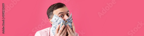 Portrait of a contented young businessman with a beard dressed in a pink jacket showing us dollar banknotes against a red studio background. Taste, smell of money.