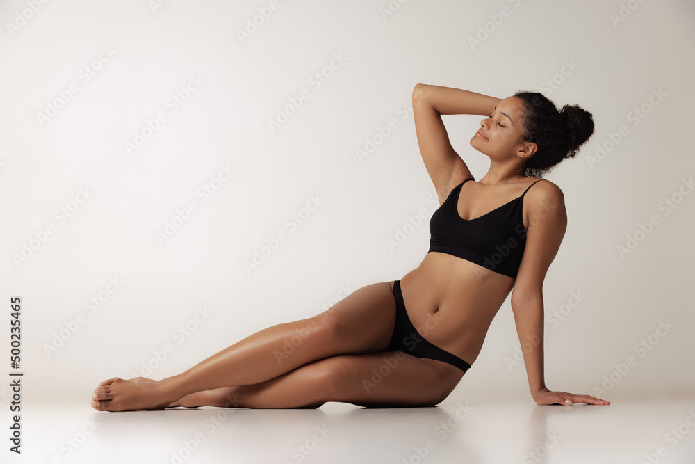 Portrait of young beautiful woman sitting on floor, posing in black underwear isolated over gray background. Perfect body