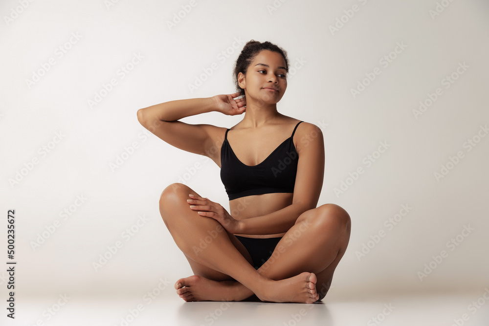 Fototapeta premium Portrait of young beautiful woman sitting on floor, posing in black underwear isolated over gray backgoround. Female health care