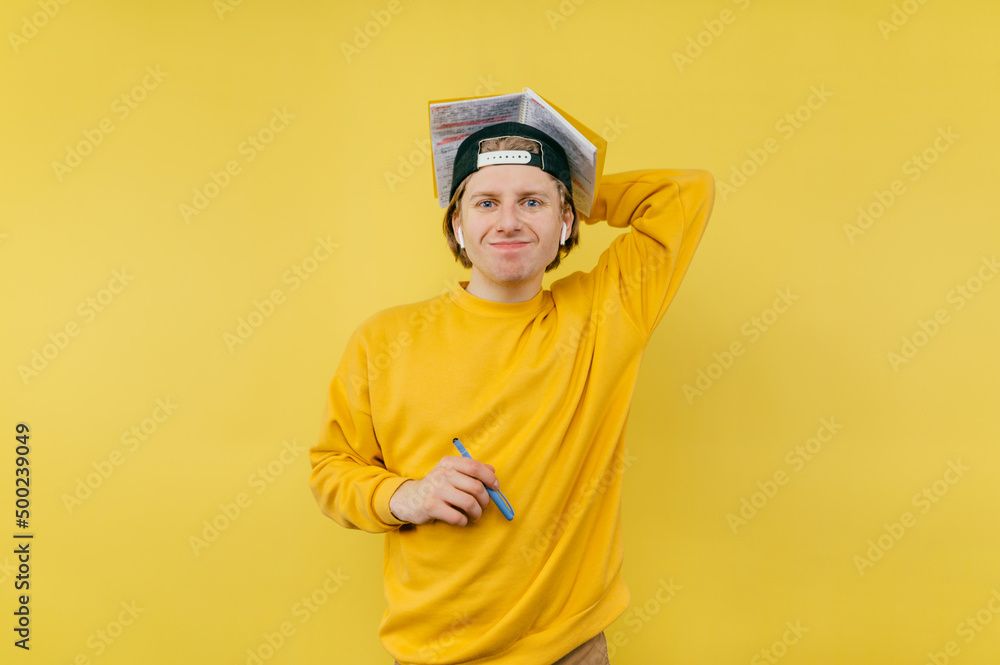 Positive young male student in casual clothes and cap standing with notebook in hand and pen, looking at camera and smiling ironically Positive young male student in casual clothes and cap standing with notebook in hand and pen, looking at camera and smiling ironically