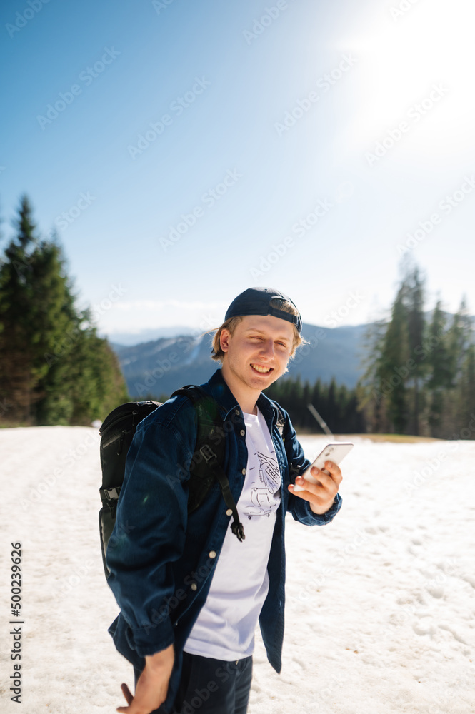 Positive young male tourist in casual clothes standing in the snow in the mountains while walking with a smartphone in his hands and looking at the camera with a smile on his face