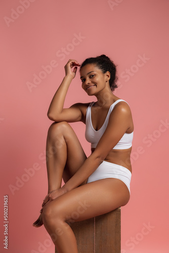 Portrait of young beautiful slim woman sitting, posing in white cotton underwear isolated over pink studio background