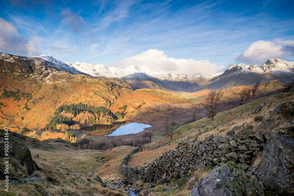 Winter landscape in the British Lake District with beautiful sunlight and snow topped mountains, looking down a stone wall to a tarn in the valley