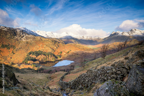 Winter landscape in the British Lake District with beautiful sunlight and snow topped mountains, looking down a stone wall to a tarn in the valley